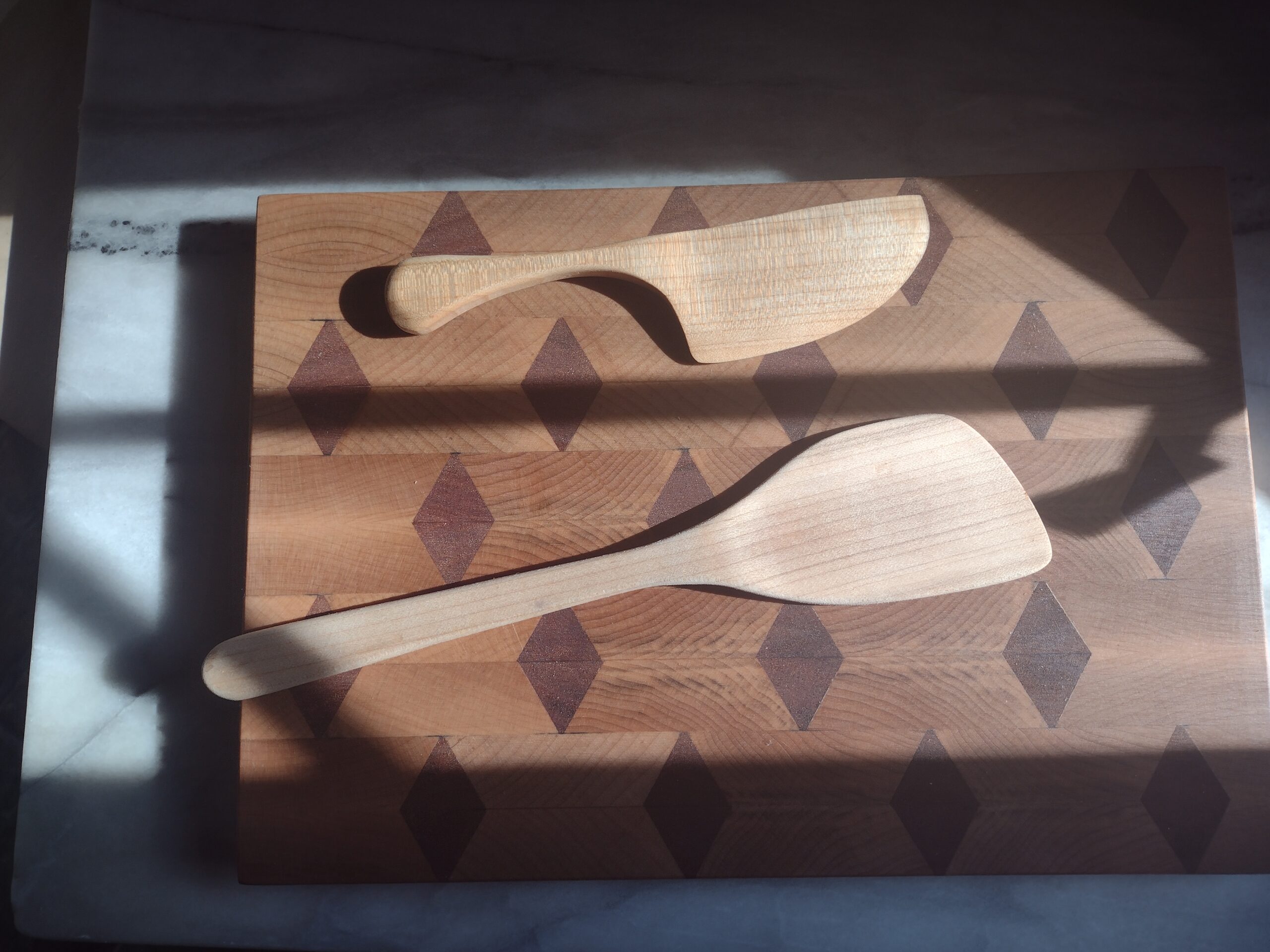 Two maple kitchen utensils: a large knife and a spatula.  Both are on a diamond-patterned cutting board lying on a piece of marble in the sunlight.